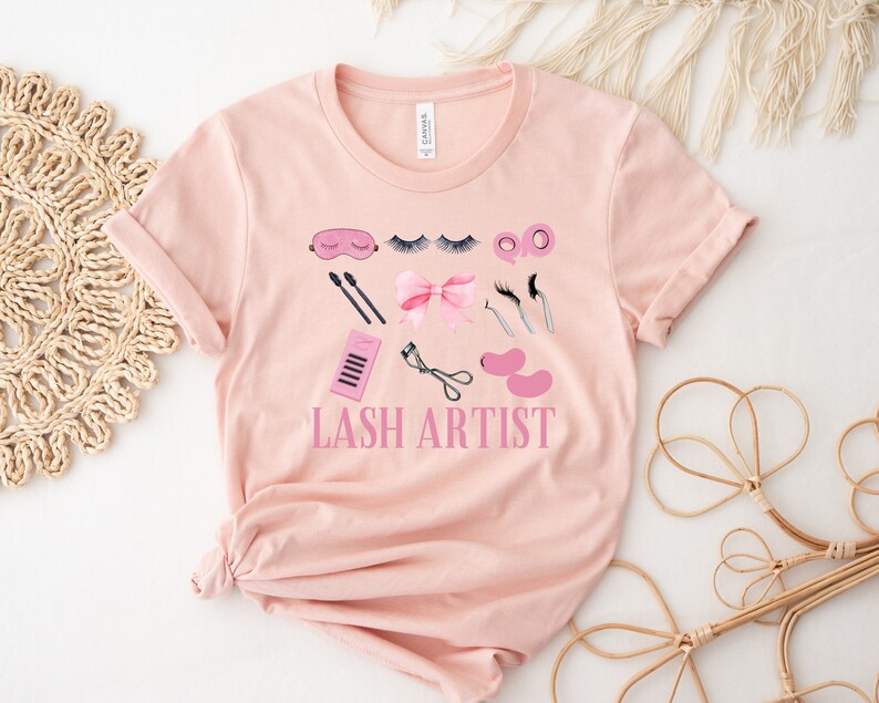 Pu&ograve; includere: Una maglietta rosa con un design grafico di strumenti per artisti di ciglia e il testo "LASH ARTIST".