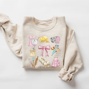 May include: A beige sweatshirt with a watercolor design of baking supplies, including a mixer, flour, eggs, a rolling pin, a bow, and a baking apron.