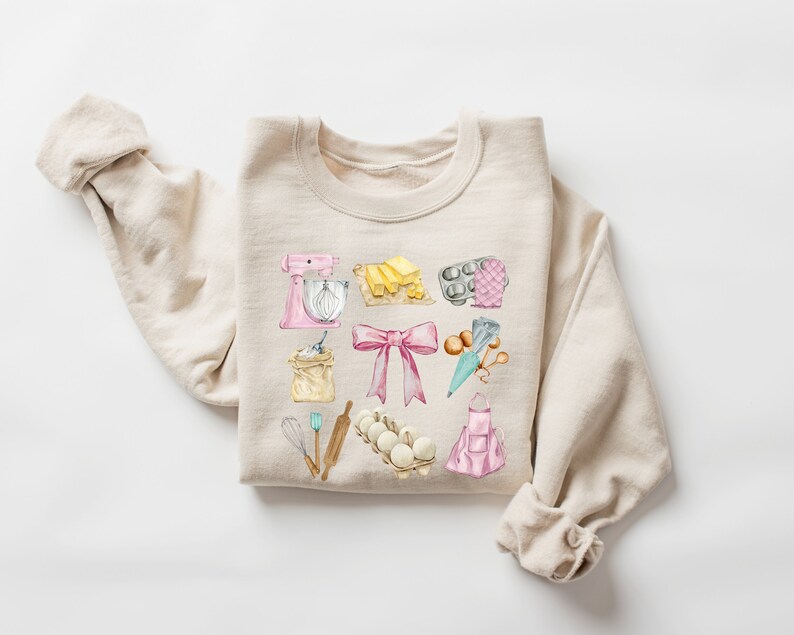 May include: A beige sweatshirt with a watercolor design of baking supplies, including a mixer, flour, eggs, a rolling pin, a bow, and a baking apron.
