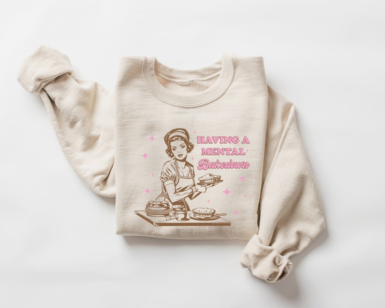 Peut inclure: Un sweat-shirt beige avec une illustration r&eacute;tro d'une femme en train de faire des tartes. Le texte "HAVING A MENTAL BAKEDOWN" est imprim&eacute; en lettres brunes et roses.