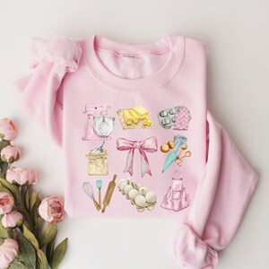 May include: Pink sweatshirt with a watercolor design of baking supplies, including a mixer, flour, butter, eggs, a rolling pin, a whisk, a piping bag, and a baking apron.