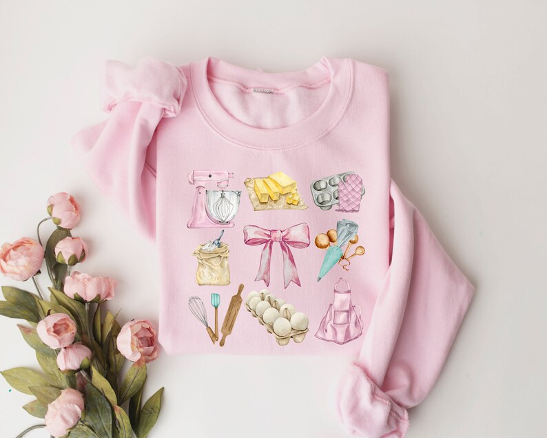 May include: Pink sweatshirt with a watercolor design of baking supplies, including a mixer, flour, butter, eggs, a rolling pin, a whisk, a piping bag, and a baking apron.