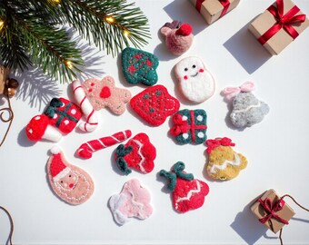 Puede incluir: Una colección de adornos navideños festivos de fieltro en varias formas y colores, incluyendo un Papá Noel, un muñeco de nieve y una caja de regalo. Los adornos están dispuestos sobre una superficie blanca con una ramita de hoja perenne y pequeñas cajas de regalo.