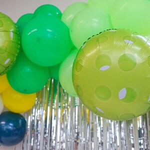 May include: A green, white, and yellow balloon arch with green polka dot balloons. The balloons are hanging from a silver tinsel curtain.