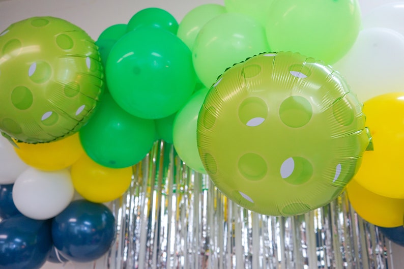 May include: A green, white, and yellow balloon arch with green polka dot balloons. The balloons are hanging from a silver tinsel curtain.