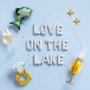 May include: A festive arrangement on a light blue background, featuring foil balloons spelling "LOVE ON THE LAKE." Includes a fish balloon, champagne glasses, a diamond ring balloon, and star-shaped balloons. Perfect for a lake-themed celebration.