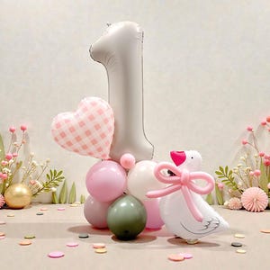May include: A celebratory balloon display for a first birthday. It includes a large silver number one balloon, a pink heart-shaped balloon, and a white goose balloon with a pink bow. The arrangement features pink, white, and green balloons.