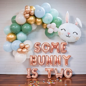 May include: A festive balloon arch in pastel colors, including white, gold, green, and blue, with a bunny-shaped balloon. Rose gold foil balloons spell out "SOME BUNNY IS TWO" against a white brick wall, perfect for a second birthday celebration.