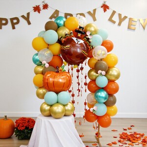 Peut inclure: Une arche de ballons festive avec des ballons orange, jaunes, dorés et turquoise, avec un ballon citrouille et dinde. Une bannière avec "HAPPY" et "LYEYEF" est suspendue au-dessus. Une citrouille et des fleurs sont au sol.