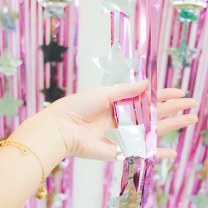 May include: A hand holds a silver and pink foil garland with star shapes. The garland is hanging against a backdrop of pink and white foil streamers.
