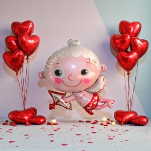 May include: A Valentine's Day-themed arrangement featuring a large Cupid-shaped balloon with a bow and arrow, surrounded by red heart-shaped balloons. The scene includes scattered rose petals and lit candles, creating a romantic atmosphere.