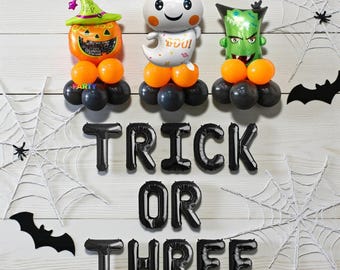 May include: Halloween-themed balloon decorations featuring a pumpkin, ghost, and Frankenstein's monster. The balloons are orange, black, and green, with the phrase "TRICK OR THREE" spelled out in black balloons. Spiderwebs and bats complete the festive scene.