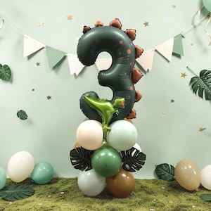 May include: A festive birthday balloon arrangement featuring a large dark green number 3 balloon with a dinosaur design. The arrangement includes a base of various colored balloons, decorative leaves, and a banner, all set against a light green backdrop.