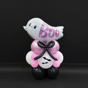 May include: A Halloween-themed balloon arrangement featuring a white ghost-shaped balloon with the word "BOO" in pink. The arrangement includes pink, white, and black balloons, with a black bow and bat silhouettes. The balloons are arranged to resemble a figure.