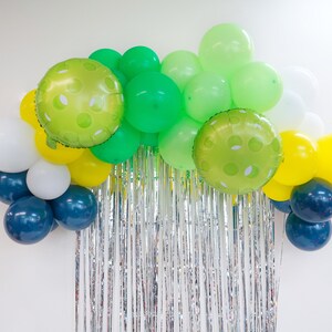 May include: A balloon arch made of green, white, yellow, and blue balloons. The balloons are arranged in a semi-circle shape and are hanging from a silver tinsel curtain.