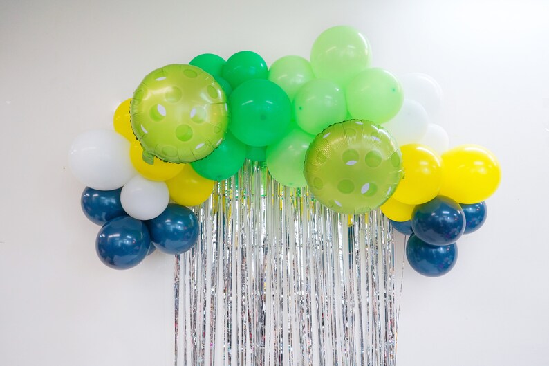 May include: A balloon arch made of green, white, yellow, and blue balloons. The balloons are arranged in a semi-circle shape and are hanging from a silver tinsel curtain.