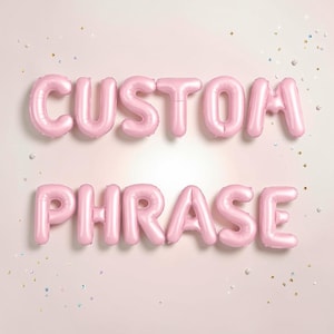 May include: Pink foil balloons spelling out "CUSTOM PHRASE" on a light pink background. The balloons are arranged in two lines, with the words clearly visible. Scattered confetti adds a festive touch to the scene.