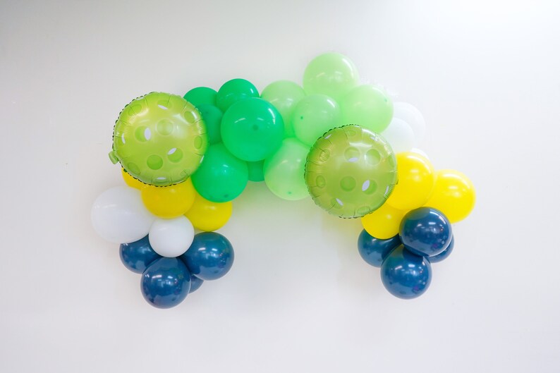 May include: A green, yellow, white, and blue balloon garland. The balloons are arranged in a semi-circle shape. The green balloons have a polka dot pattern.