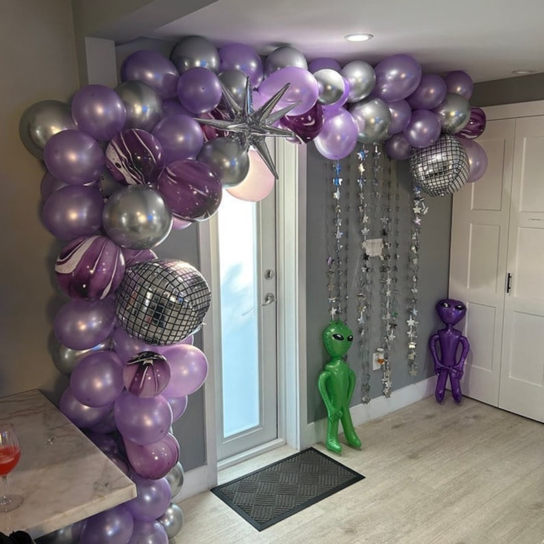 May include: A purple and silver balloon archway with disco ball decorations. Two alien figures stand in front of the archway.