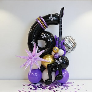 Puede incluir: Arreglo de globos negros con el número seis, con detalles morados y dorados. Incluye una guitarra morada, una bola de discoteca y notas musicales. Decoraciones de estrella y mariposa. Globos sobre una superficie blanca con confeti morado.