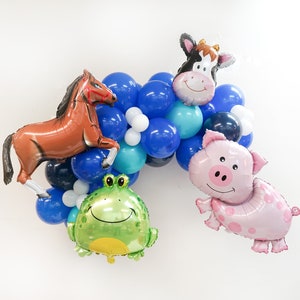 K&ouml;nnte beinhalten: Ein Ballonbogen mit blauen, wei&szlig;en und t&uuml;rkisfarbenen Ballons. Der Bogen enth&auml;lt tierf&ouml;rmige Ballons, darunter ein Pferd, eine Kuh, ein Schwein und ein Frosch.