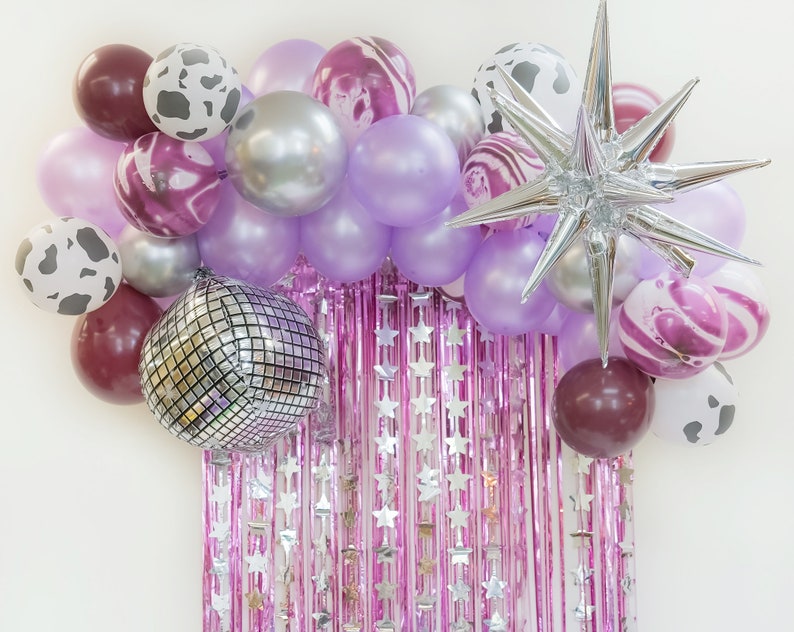 May include: A purple, pink, and silver balloon garland with a silver star balloon. The garland is decorated with cow print balloons, marble print balloons, and disco ball balloons.