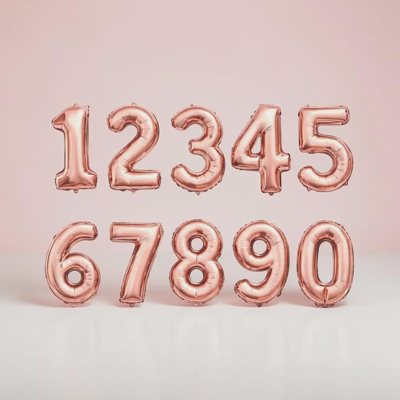 May include: Rose gold foil number balloons, including digits 0 through 9. The balloons are arranged in two rows against a soft pink background. These balloons are ideal for birthdays, anniversaries, or other celebratory events.