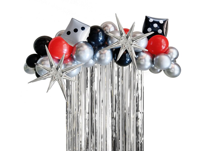 May include: A silver and black balloon arch with red balloons, silver stars, and a dice-shaped balloon. The arch is decorated with silver tinsel streamers.