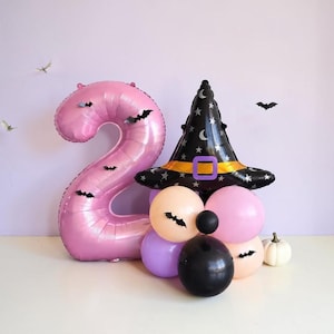 May include: A large pink number two balloon decorated with small black bat cutouts, paired with a black witch hat balloon resting on a cluster of round peach, pink, purple, and black balloons, with a small white pumpkin nearby.