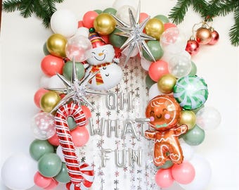 Könnte beinhalten: Ein festlicher Weihnachtsballonbogen mit Schneemann-, Lebkuchenmann- und Zuckerstangenballons. Der Bogen enthält goldene, weiße, rosa und grüne Luftballons mit silbernen Sternen. Der Text "OH WHAT FUN" wird in silbernen Luftballons angezeigt.