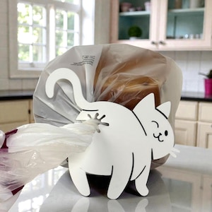 Puede incluir: Un soporte para bolsas con forma de gato blanco hecho de metal. El gato tiene una cola curvada y está diseñado para sujetar bolsas de plástico. El soporte está sobre una encimera en un entorno de cocina, con una bolsa de pan al fondo.