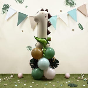 May include: A festive balloon arrangement for a first birthday celebration. The centerpiece is a large number one balloon, with a dinosaur-shaped balloon and a cluster of green, brown, and white balloons. The background includes a banner and decorative elements.