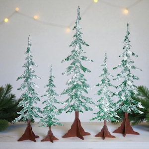 May include: A set of five decorative Christmas tree figurines. The trees are green with white accents, resembling snow. They have brown bases and are arranged in a row. The background is a light color with a string of lights.