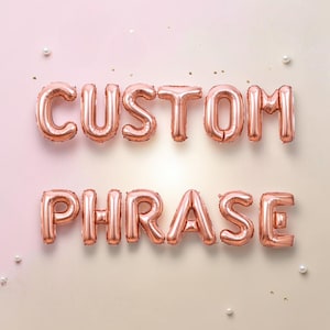 May include: Rose gold foil balloons spelling out "CUSTOM PHRASE" against a light pink background. The balloons are arranged in two lines, with the word "CUSTOM" above "PHRASE". Scattered confetti and pearls add a festive touch.