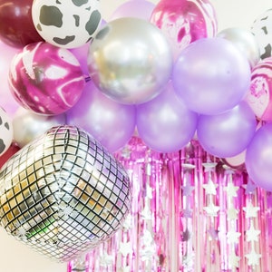 May include: A balloon arch with a mix of purple, silver, and pink balloons. The balloons are arranged in a semi-circle shape and are decorated with silver glitter and white polka dots.