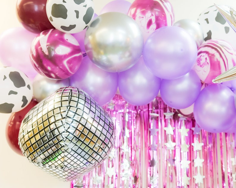 May include: A balloon arch with a mix of purple, silver, and pink balloons. The balloons are arranged in a semi-circle shape and are decorated with silver glitter and white polka dots.