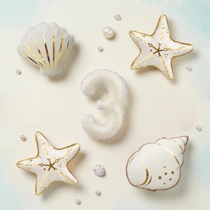 May include: A set of white and gold foil balloons featuring a number three, two starfish, and two seashells, accented by scattered white pearl-like spheres. The balloons have a metallic gold printed pattern on a matte white surface.