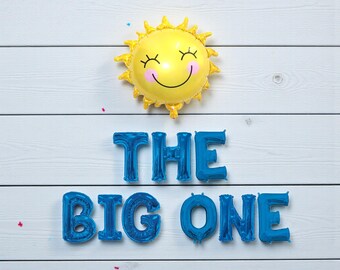 May include: A yellow sun-shaped balloon with a smiling face and the text "THE BIG ONE" in blue balloon letters, set against a white wooden background. Confetti is scattered around the balloons.