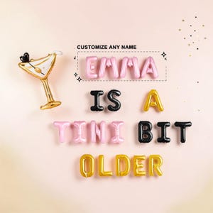 May include: A set of foil letter balloons spelling out EMMA IS A TINI BIT OLDER, accompanied by a martini glass-shaped balloon. The text CUSTOMIZE ANY NAME is printed above the name EMMA. The balloons are in shades of pink, black, and gold.
