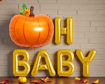 Puede incluir: Un globo con forma de calabaza naranja con detalles verdes y amarillos, y globos con letras doradas que deletrean "OH BABY". Una pequeña calabaza y hojas de otoño sobre una superficie de madera.