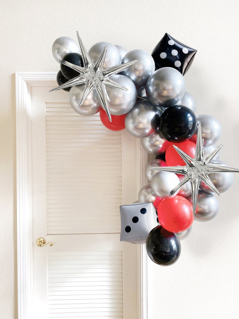 May include: A silver, black, and red balloon arch with star-shaped balloons and dice-shaped balloons. The arch is decorated with black and white polka dots.
