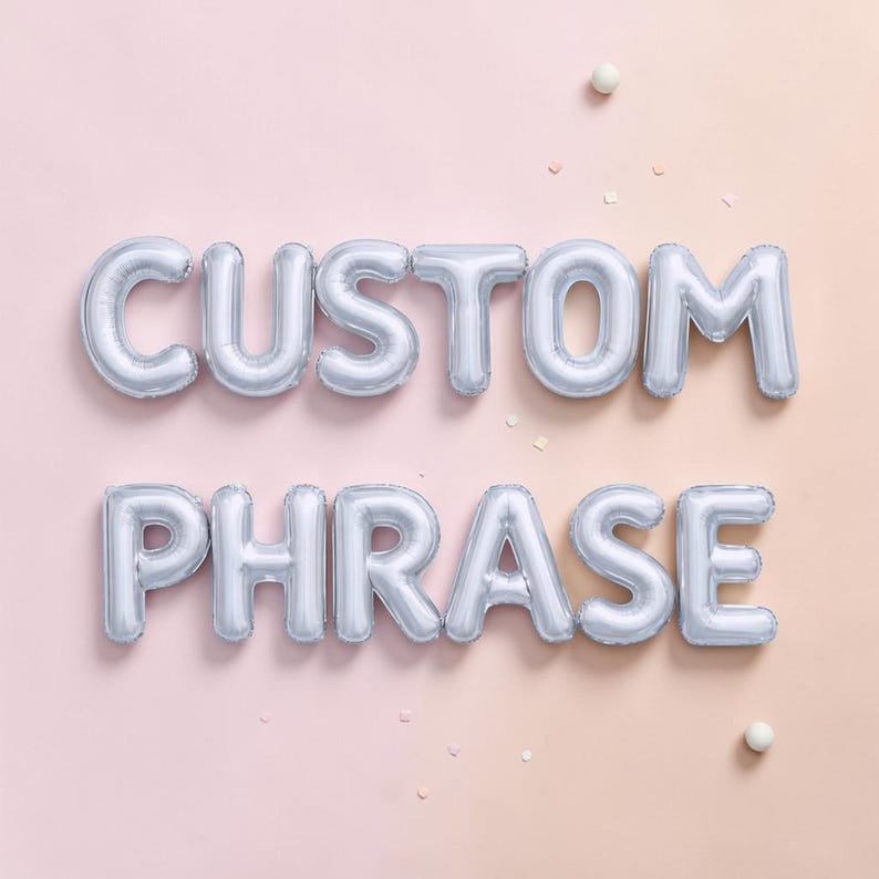 May include: Silver foil balloons spelling out "CUSTOM PHRASE" against a soft pink background. The balloons are inflated and arranged in two rows. Small white confetti and spheres are scattered around the balloons.