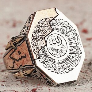 May include: A silver-toned metal ring featuring an octagonal face engraved with intricate Arabic calligraphy. The sides of the ring display additional etched script and decorative metalwork, showcasing a polished finish with dark recessed details.
