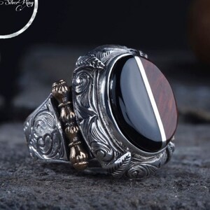 Può includere: Un anello d'argento con una pietra ovale di onice nero incastonata in una fascia d'argento dettagliata. La fascia presenta intricati disegni floreali e un piccolo accento dorato.
