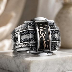 May include: A silver ring featuring a rectangular center panel with engraved Arabic calligraphy in gold tone, surrounded by rows of small black stones. The band has a textured, oxidized finish with additional black stone accents on the sides.