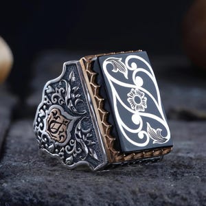 May include: A rectangular silver ring featuring a black stone inlay with an engraved white floral pattern. The silver band is decorated with intricate scrollwork and a small gold-toned accent on the side, showcasing detailed metal craftsmanship.