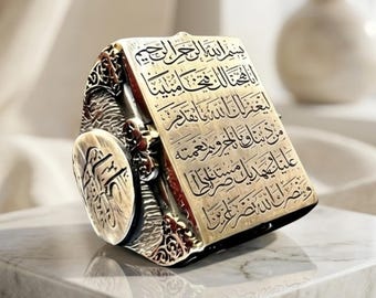 May include: A silver ring featuring a rectangular face engraved with Arabic calligraphy and intricate side detailing. The band displays a circular emblem on the side, with the text RS Reyhan Silver visible at the bottom of the frame.