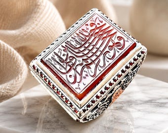 May include: A rectangular silver ring featuring a deep red stone inlay with intricate Arabic calligraphy engraved on the surface. The sides of the ring band are decorated with detailed black and silver filigree patterns and small red accents.