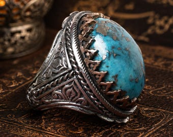 May include: A silver ring featuring a large, oval-shaped turquoise stone with dark matrix inclusions. The band is crafted with intricate, embossed floral and geometric patterns, and the stone is secured by a serrated metal setting. Text reads Reyhan Silver.