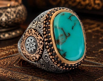 May include: A silver ring featuring a large oval turquoise stone with brown veining, surrounded by a border of small black stones. The band displays intricate scrollwork, gold-toned accents, and a central floral motif on the side of the metal setting.